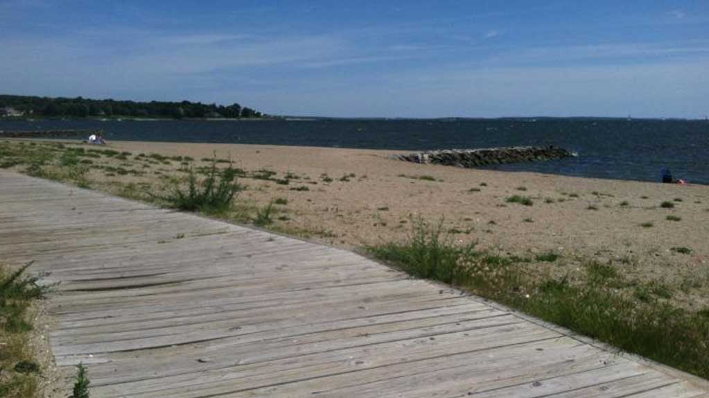 Oakland Beach in Warwick, RI, featuring sandy shores along Narragansett Bay, gentle waves, boats on the water, and a clear sky.