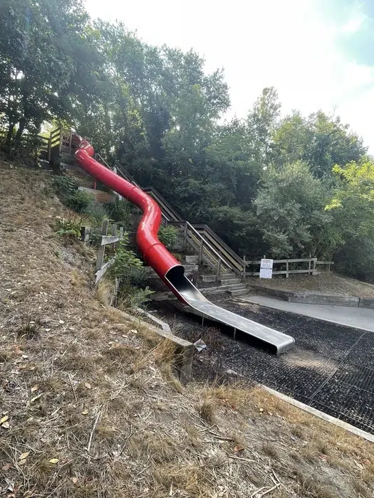 A large red slide at Pierce Beach Park in Somerset, MA, surrounded by a spacious playground area, lush green trees, and a bright blue sky, within Pro Mosquito Solutions service area.