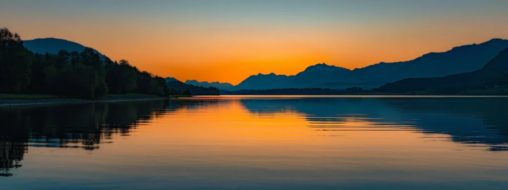 a vibrant sunset casts golden hues over a serene lake, reflecting the majestic silhouette of distant mountains under a clear, twilight sky.